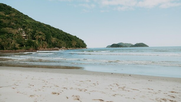 A beach with white sand, calm water, and a forested hillside. - Drifter Hostel (São Sebastião)