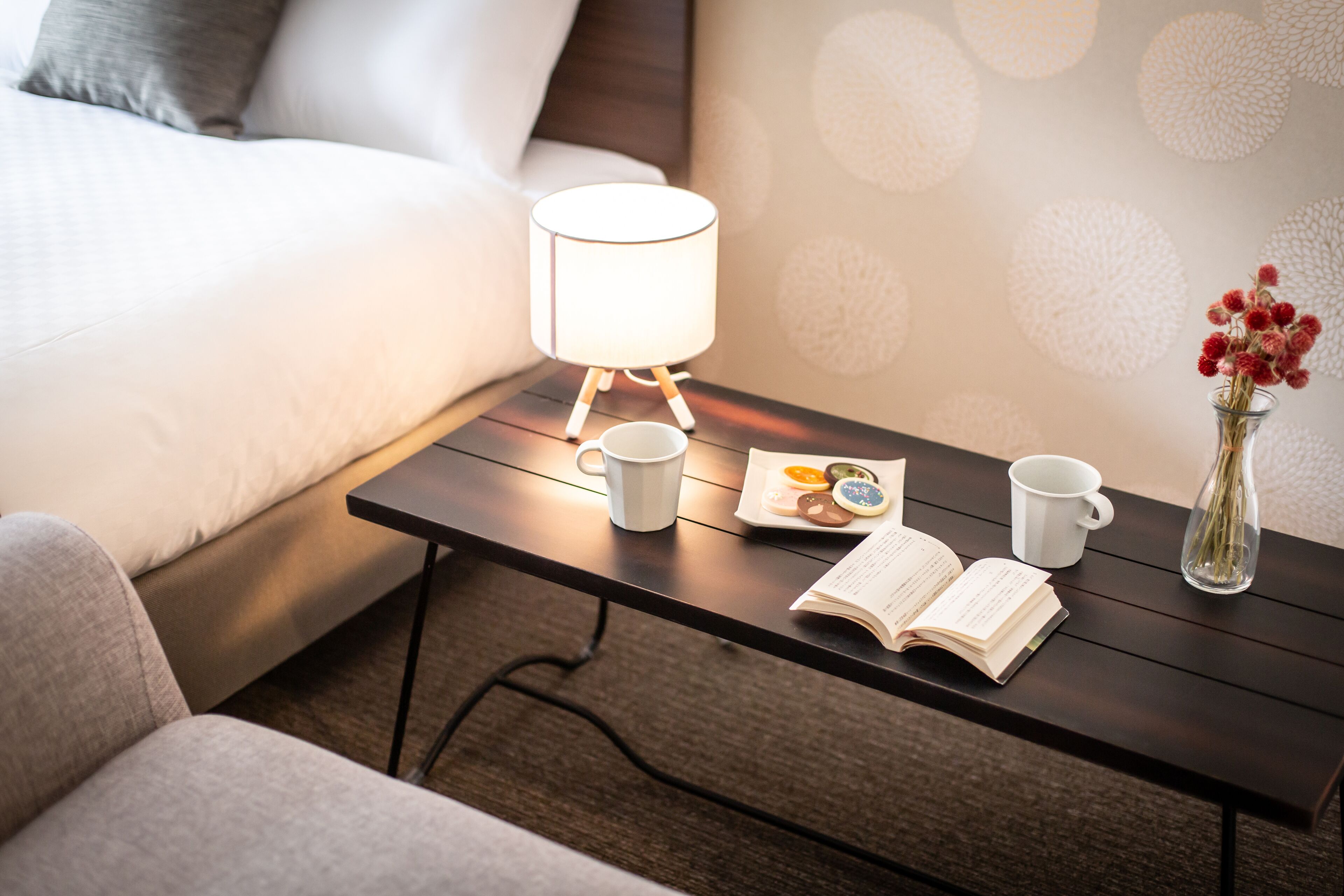 A neatly arranged hotel room with a bed, a bedside table with a lamp, a cup, a book, and a vase with flowers.