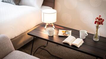 A neatly arranged hotel room with a bed, a bedside table with a lamp, a cup, a book, and a vase with flowers.