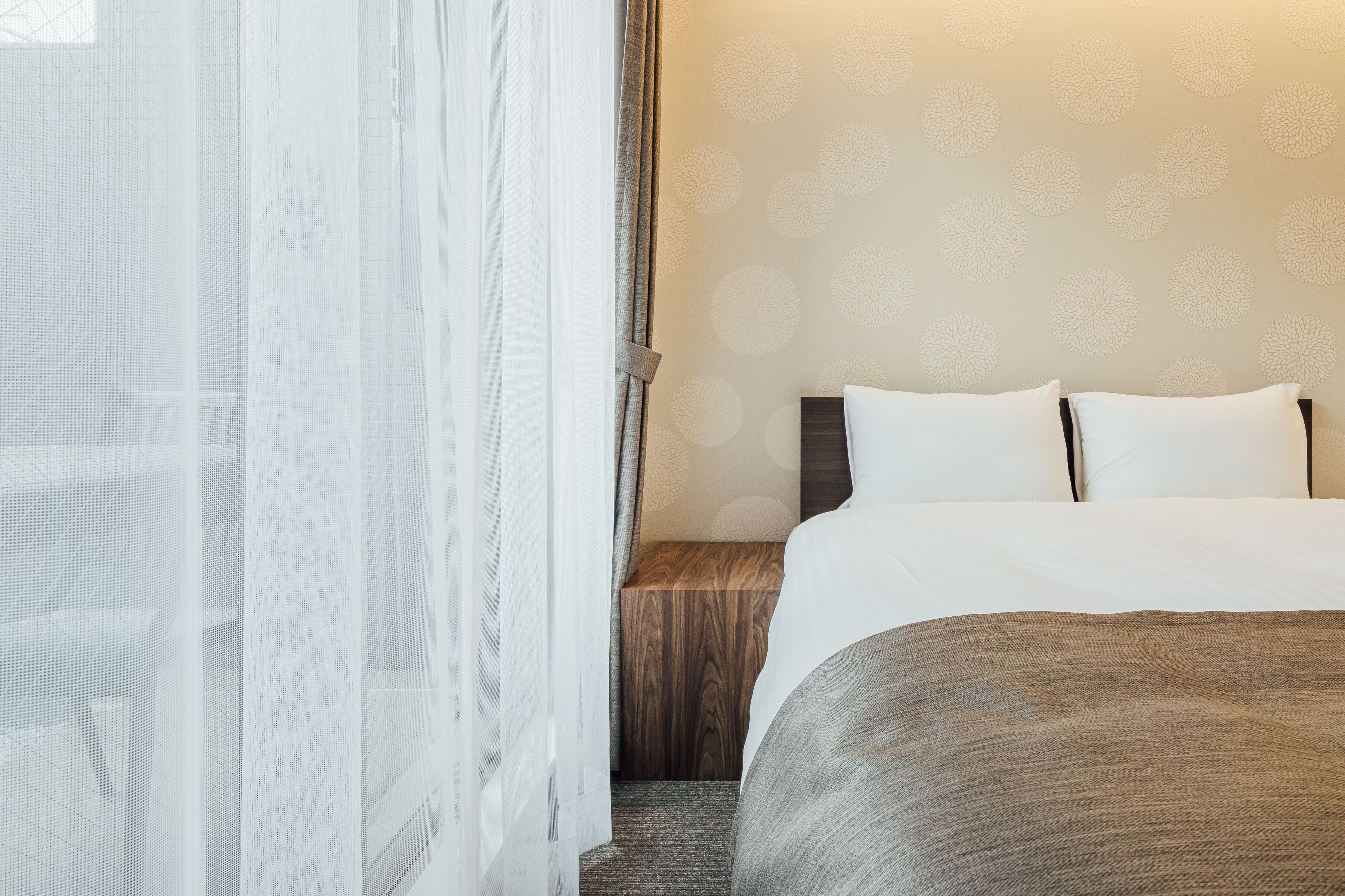 A hotel room with a bed, a wooden nightstand, and sheer curtains.