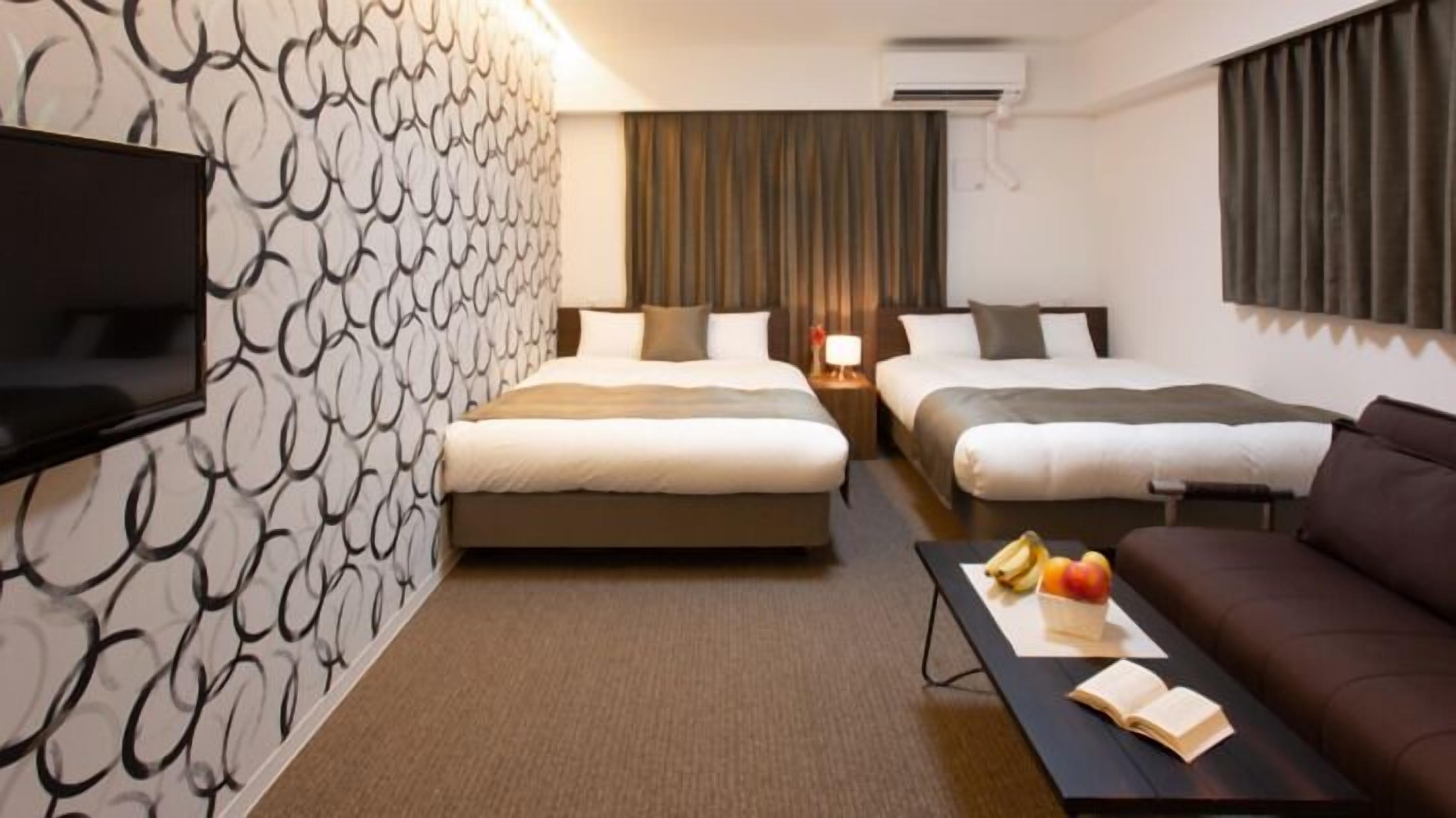 A hotel room with two beds, a TV, a sofa, a table with fruits, and a patterned wall.