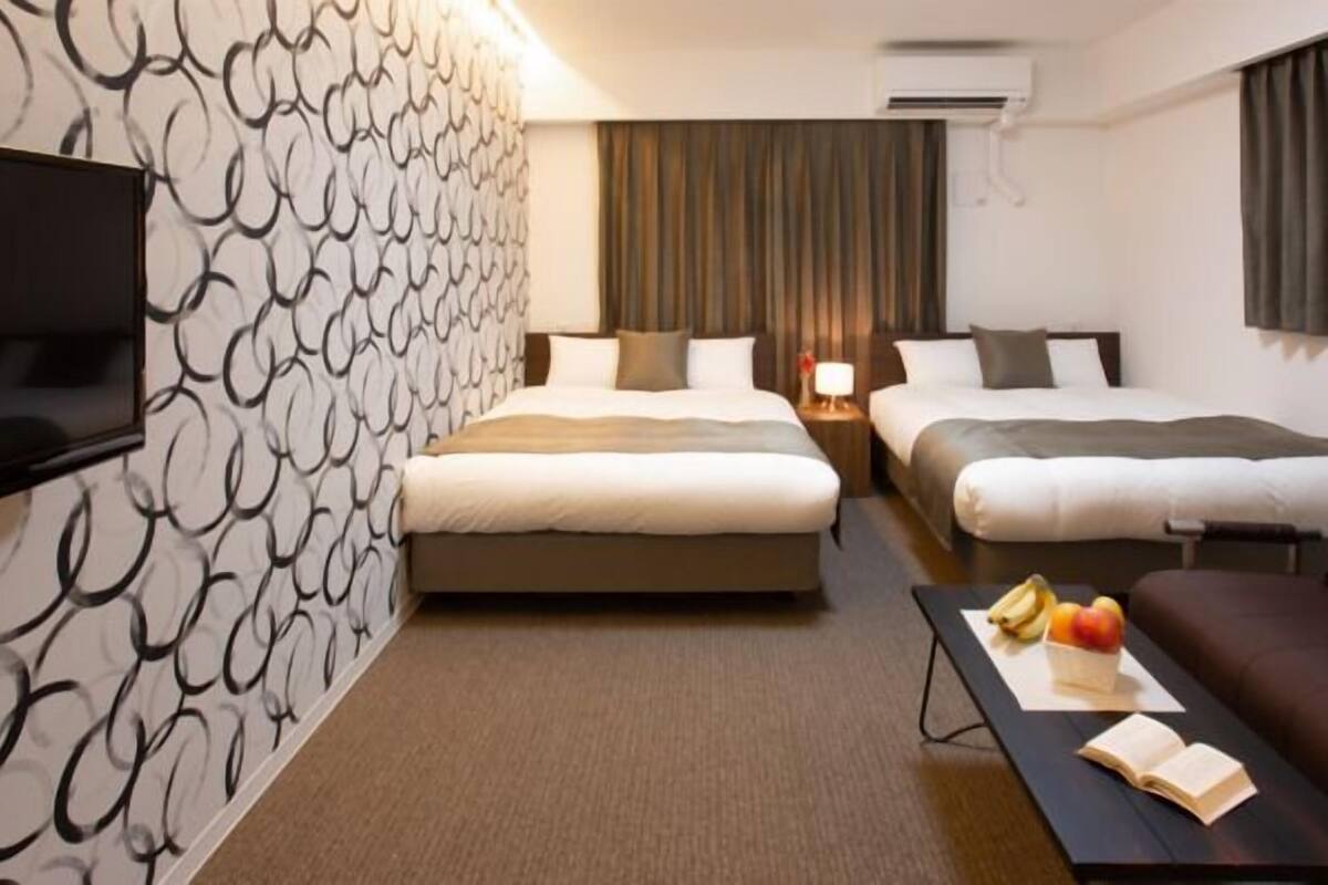 A hotel room with two beds, a TV, a sofa, a table with fruits, and a patterned wall.