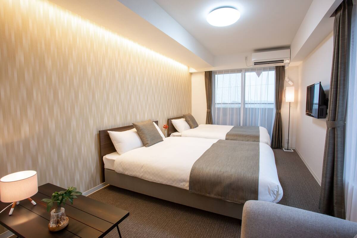 A hotel room with two beds, a desk with a lamp, a plant, and a TV on the wall.