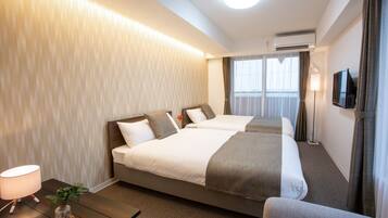 A hotel room with two beds, a desk with a lamp, a plant, and a TV on the wall.