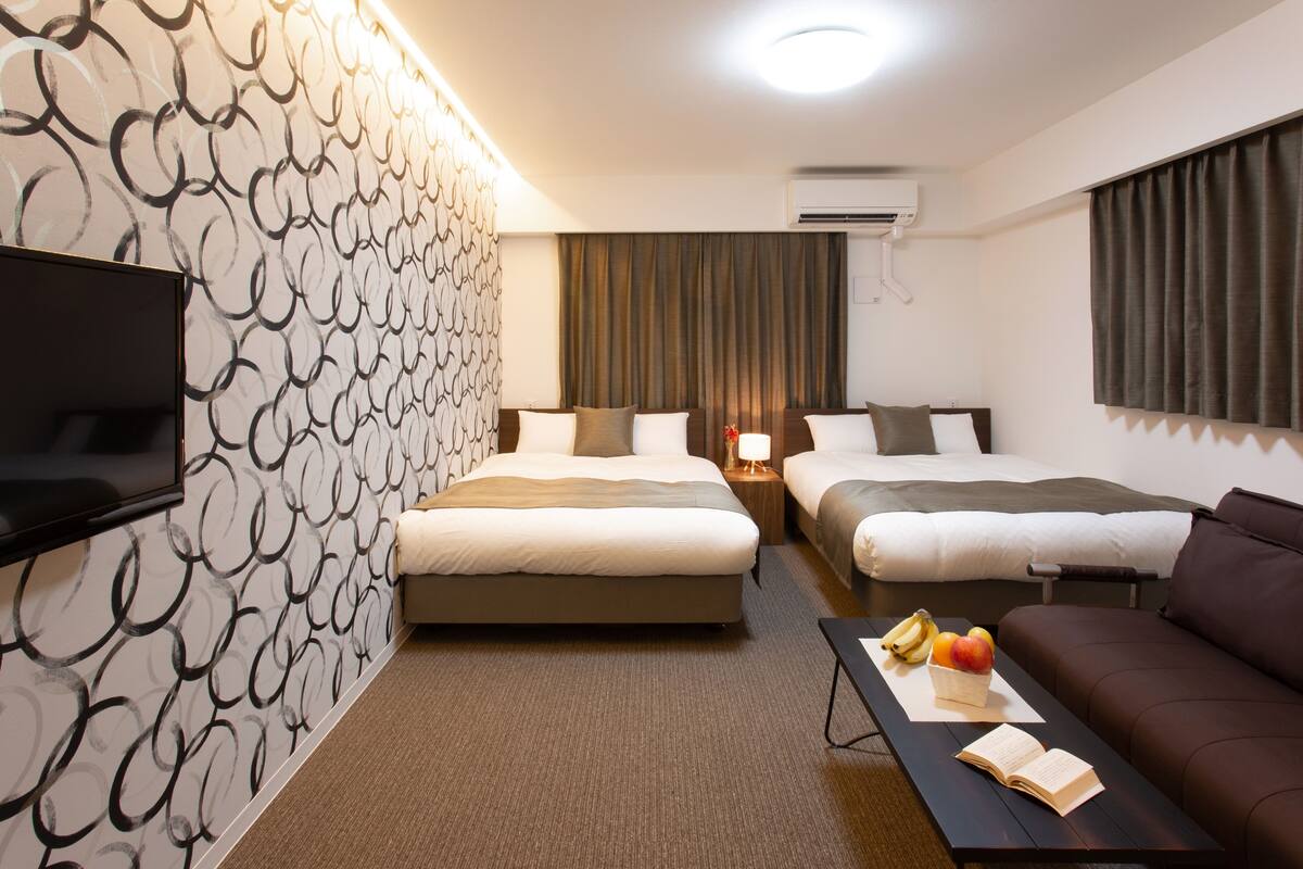 A hotel room with two beds, a sofa, a TV, and a table with fruit and a book.
