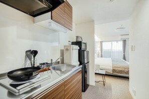 Cozinha compacta com fogão, pia e geladeira, que dá acesso a um quarto com cama e janela.