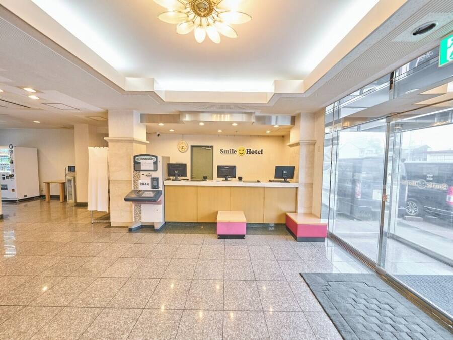 A hotel reception area with a reception desk, a digital check-in kiosk, and a large window offering a view of the street outside.