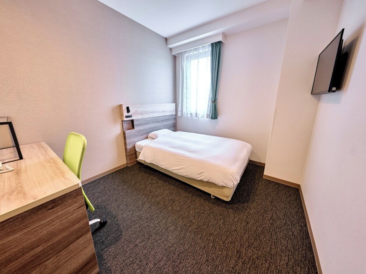 A small hotel room with a single bed, a desk, a chair, a TV mounted on the wall, and a window with curtains.