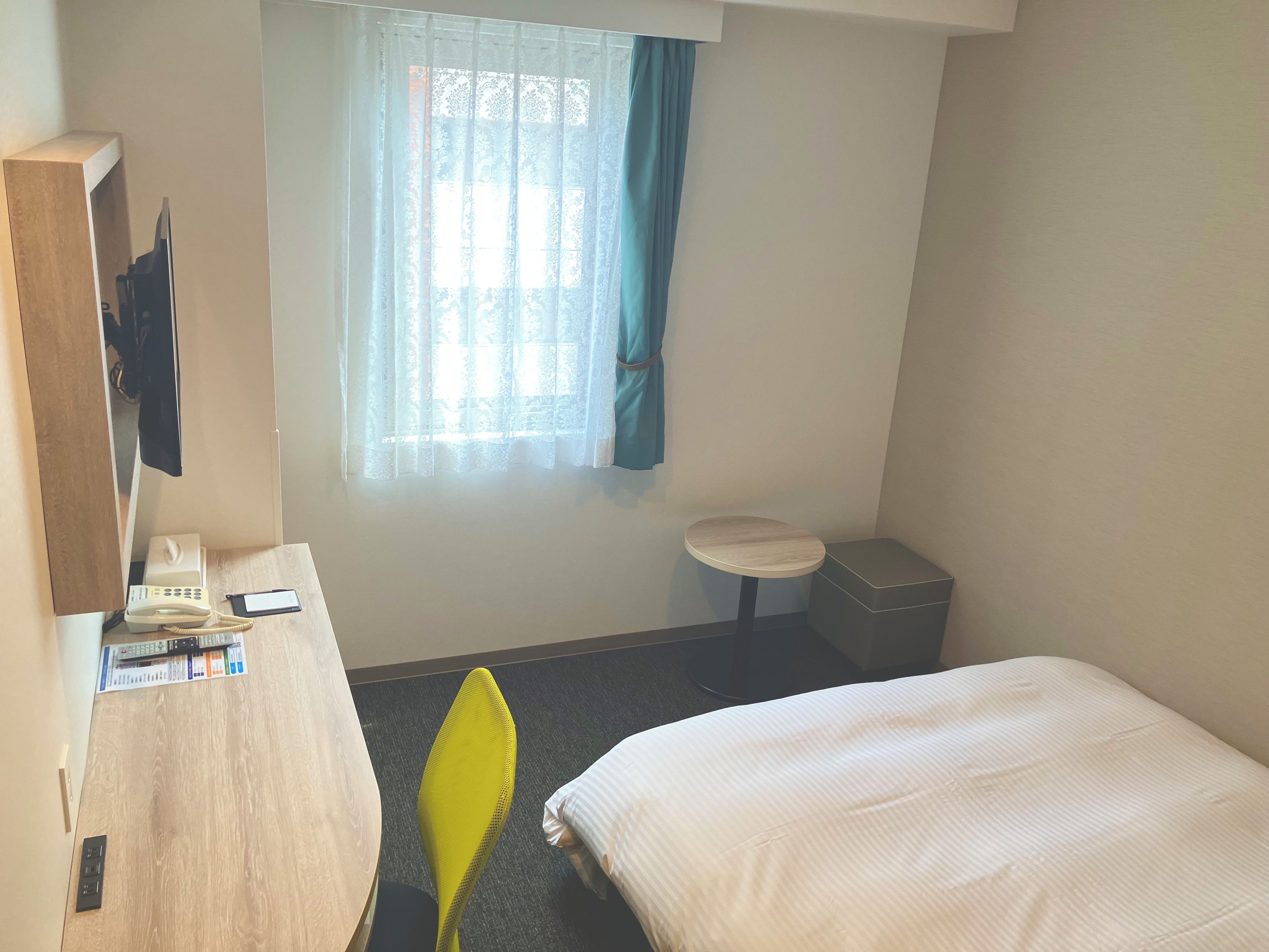 A hotel room with a bed, a desk with a telephone and papers, a small table, a trash bin, and a window with curtains.