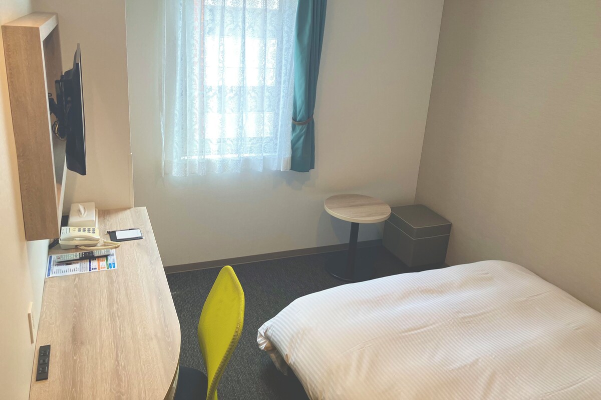 A hotel room with a bed, a desk with a telephone and papers, a small table, a trash bin, and a window with curtains.