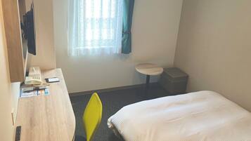 A hotel room with a bed, a desk with a telephone and papers, a small table, a trash bin, and a window with curtains.