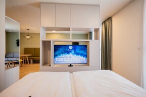 A modern hotel room with a large bed, a flat-screen TV mounted on the wall, a small seating area with a table and chairs, and a view of the room's interior.
