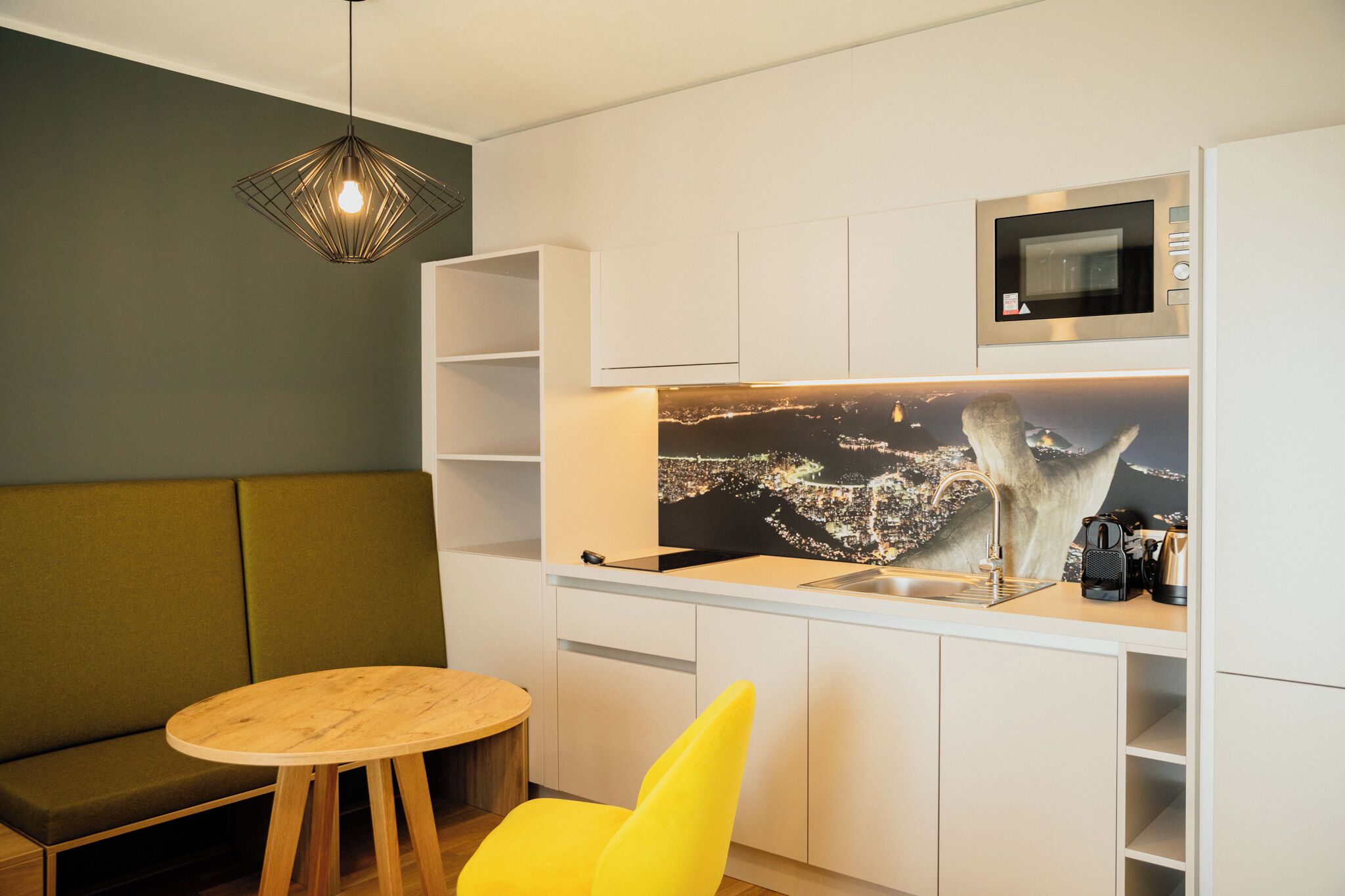 A compact kitchenette with a microwave, a sink, and a cityscape mural. There is a round wooden table and a yellow chair.