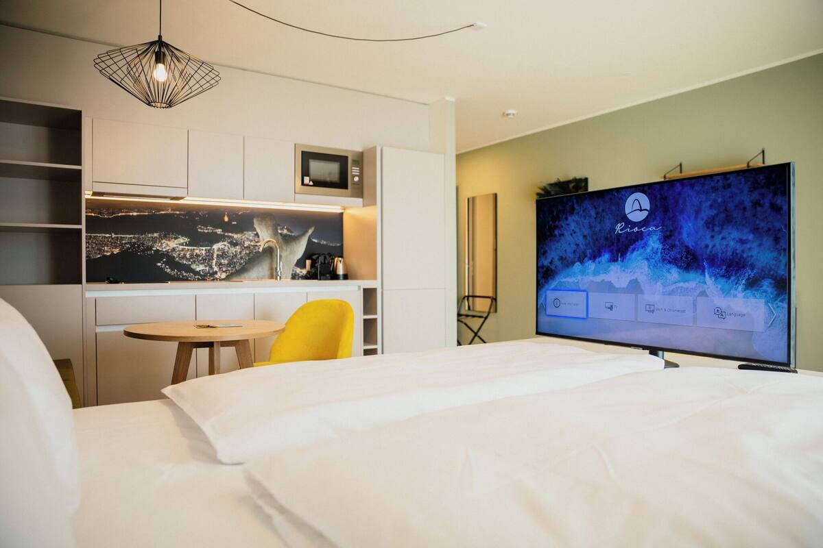 Habitación de hotel con cama, escritorio con silla, televisor de pantalla plana y vista panorámica de la ciudad en la pared.