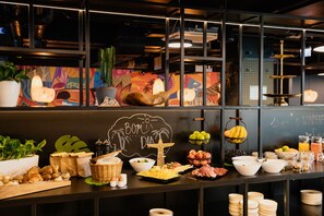 A buffet setup with various food items, a chalkboard with 'BOM DIA' written on it, and a decorative cross.