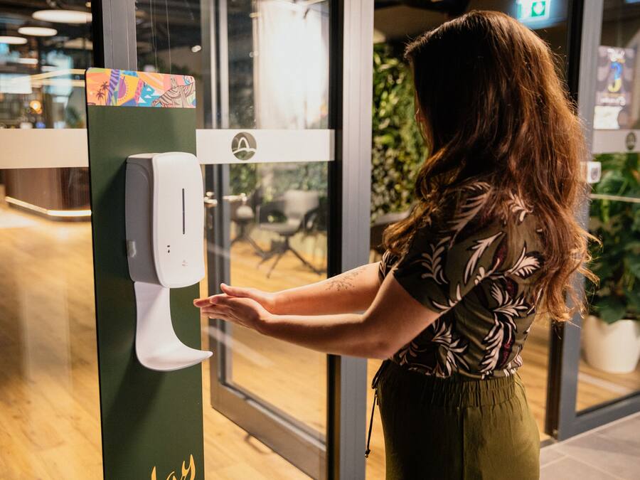 A person using a hand sanitizer dispenser in a public indoor space with a view of the outdoors through glass doors.
