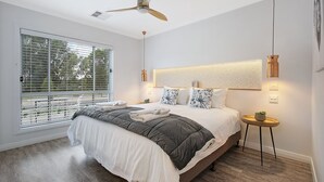 Un dormitorio con una cama grande, un ventilador de techo, una ventana con persianas y una mesita de noche con una planta.