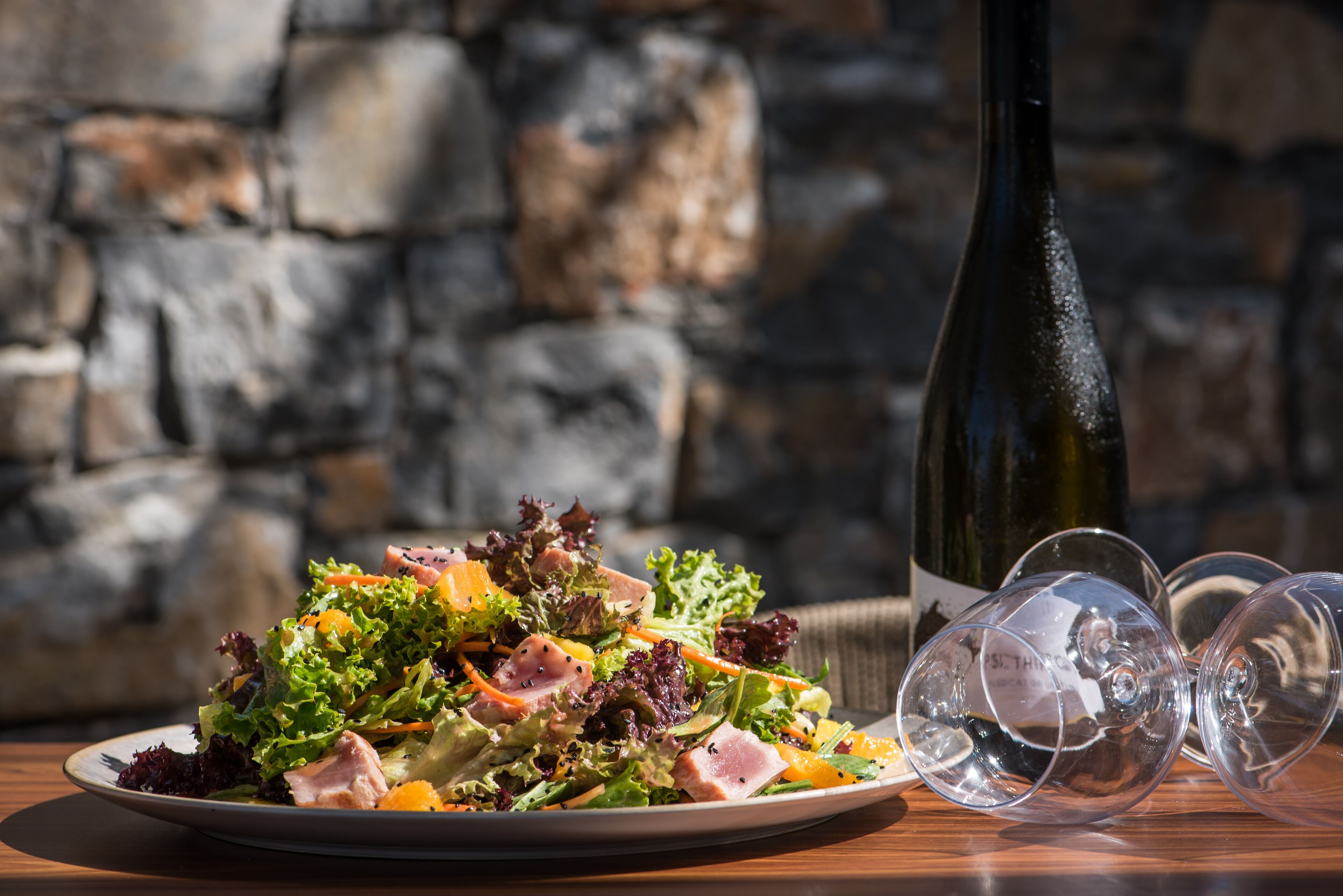 Un plato de ensalada con diferentes verduras y carnes, una botella de vino y dos copas de vino sobre una mesa de madera con un fondo de pared de piedra.