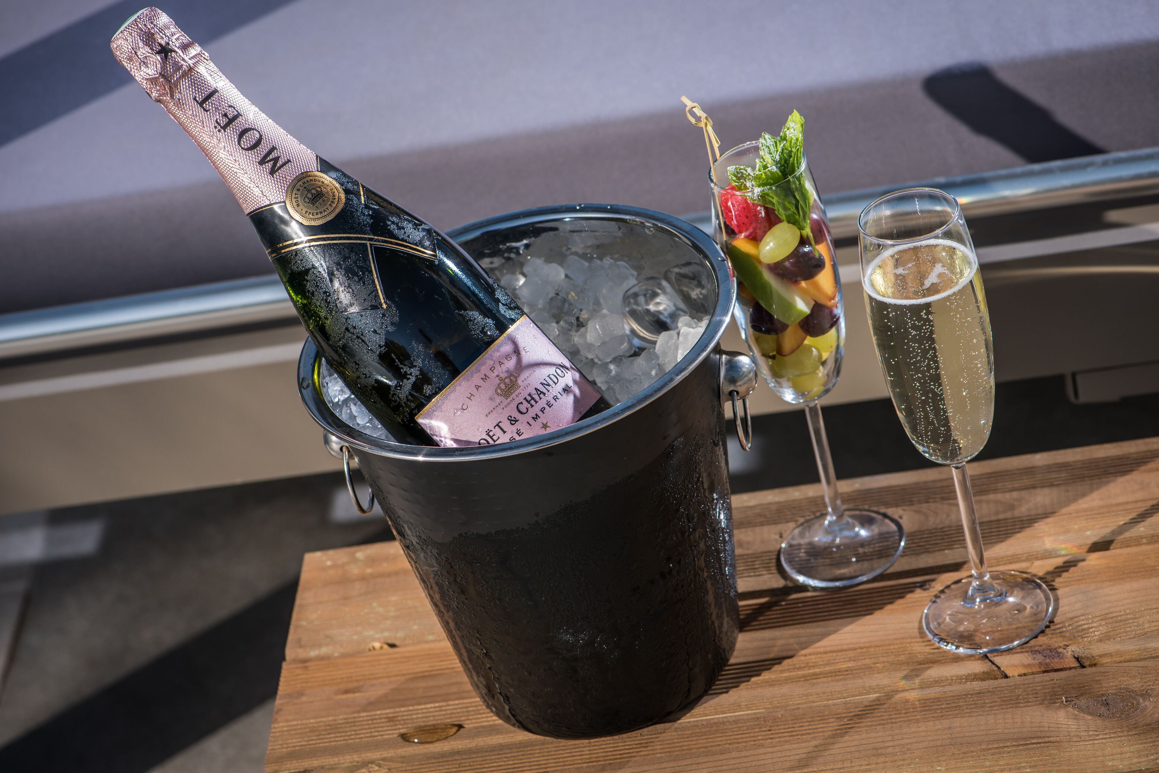 Eine Champagnerflasche in einem Eiskübel, ein Obstcocktail im Glas und ein mit Champagner gefülltes Glas auf einem Holztisch.