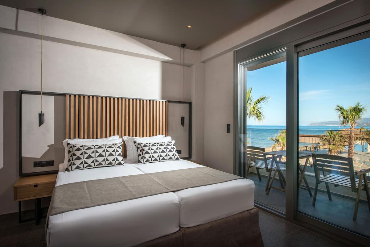 A modern hotel room with a large bed, a wooden headboard, and a balcony with a view of the sea and palm trees.