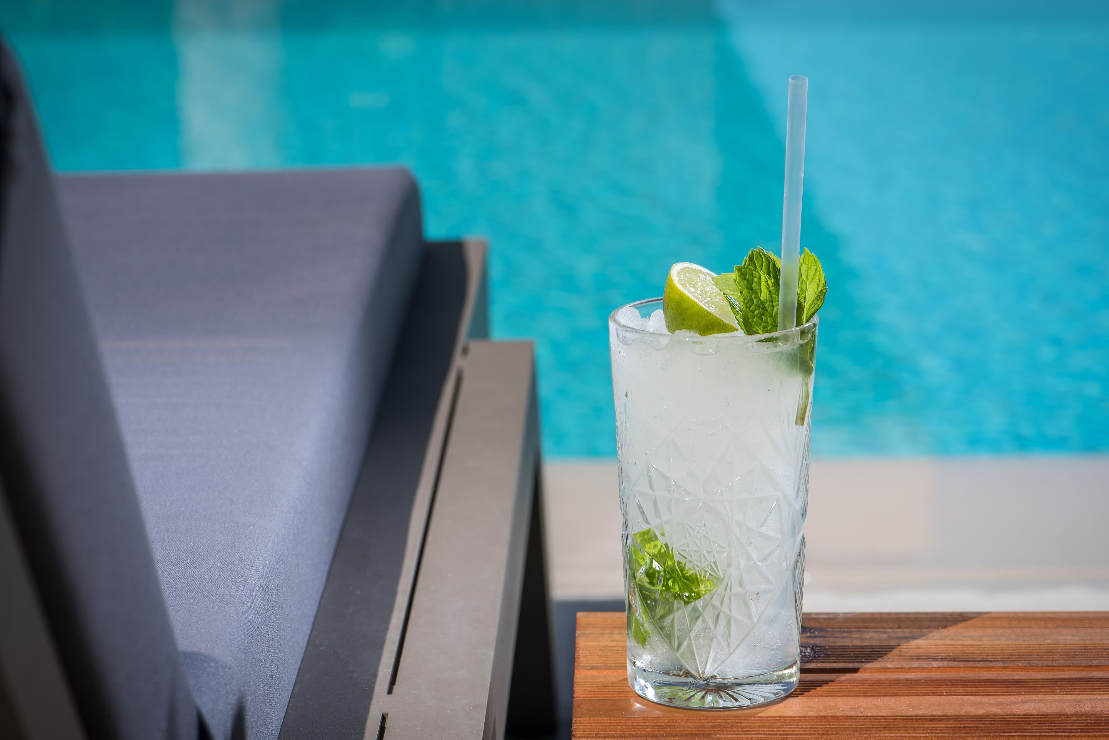 A glass of a refreshing beverage with mint and lime, placed on a wooden table by a poolside lounge chair.