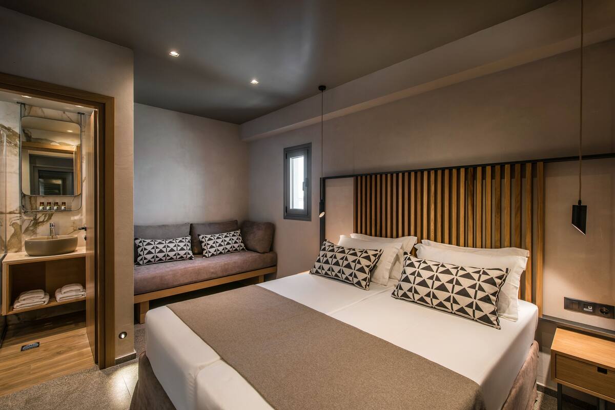 A modern bedroom with a large bed, a wooden headboard, a bedside table, a built-in bench with cushions, a large mirror, and a vanity with a sink.