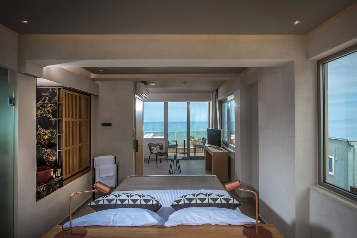 A modern hotel room with a large bed, a desk, and a balcony with a view of the sea.