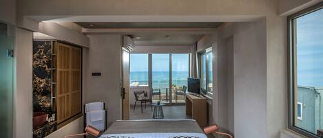 A modern hotel room with a large bed, a desk, and a balcony with a view of the sea.