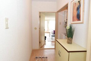 Un pasillo con piso de madera que lleva a un dormitorio con cama y ventana. Hay un pequeño armario con un jarrón de flores encima.