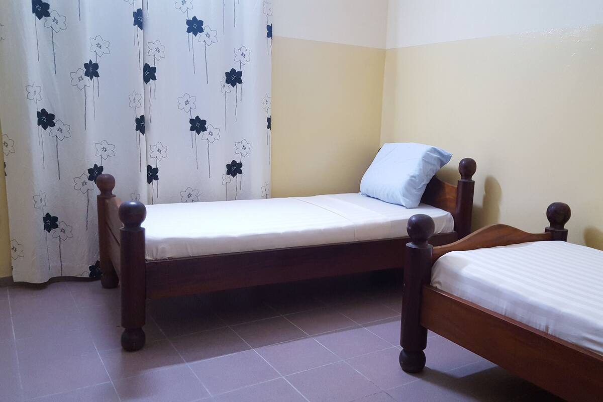 A room with two wooden beds, a white curtain with a floral pattern, and a plain yellow wall.