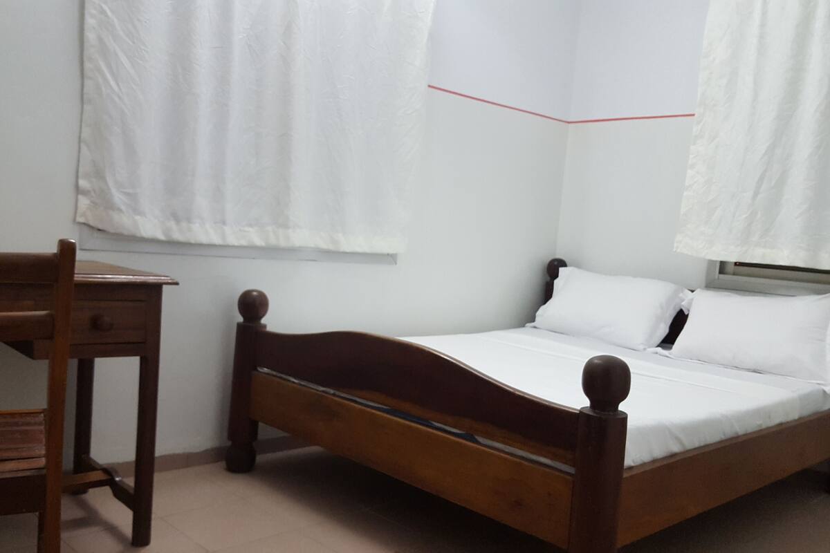 A bedroom with a wooden bed, a wooden chair, and a window with white curtains.