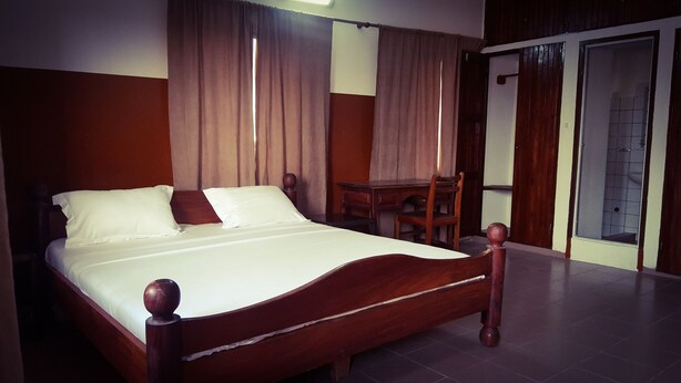 A neatly made bed with white linens, a wooden headboard, and a wooden nightstand with a lamp. - Academia Hotel (Lomé)