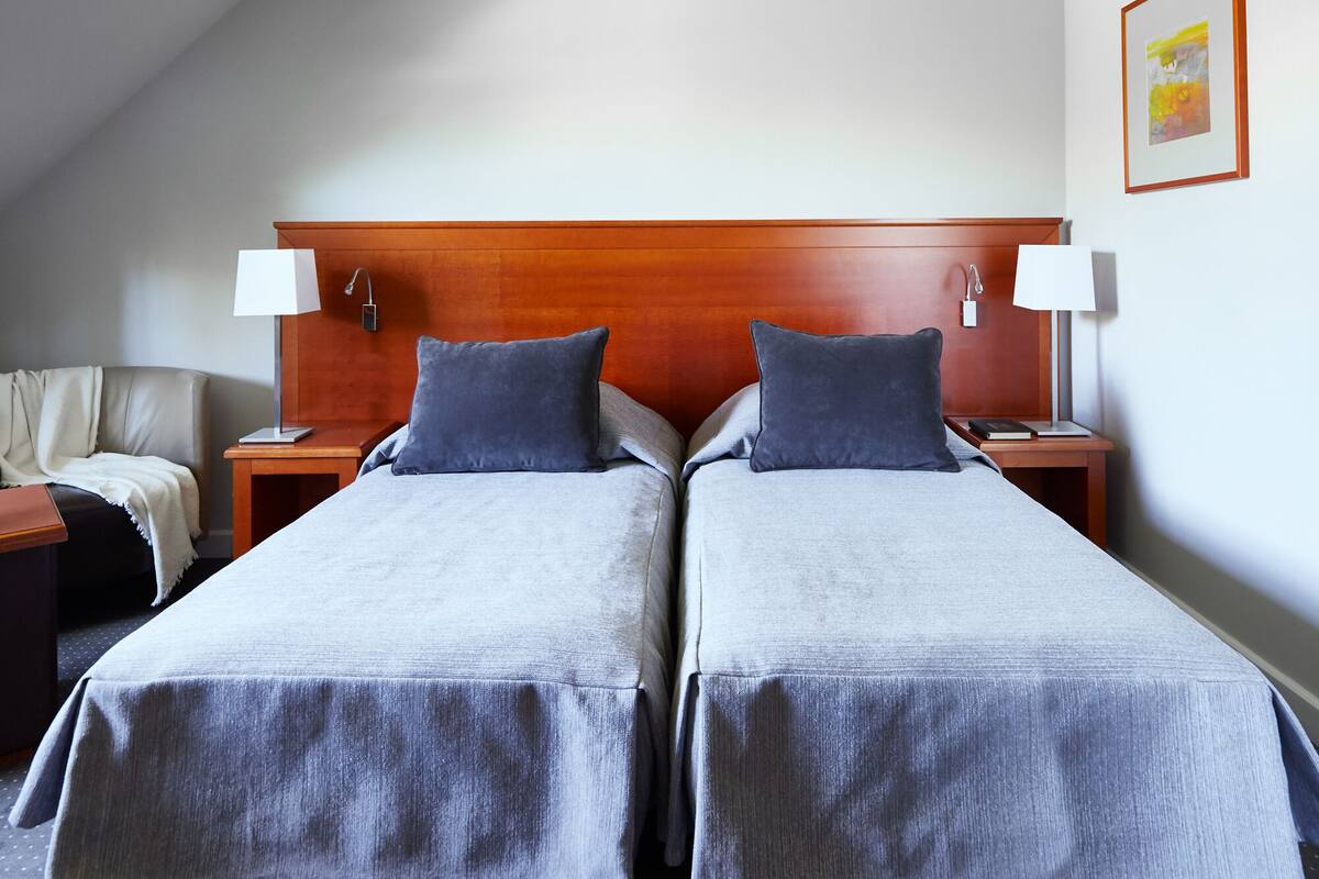 Habitación de hotel con dos camas, cabecera de madera, mesitas de noche, lámparas y un cuadro en la pared.