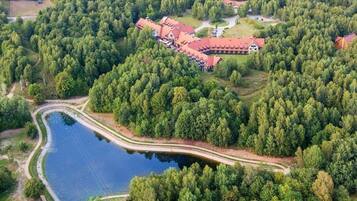 An aerial view of a lodge surrounded by dense forest, with a lake and a winding road.