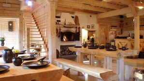 Interior rústic de fusta amb una llar de foc, una escala de fusta i una zona de dinar amb bancs i una taula preparada per a menjar.