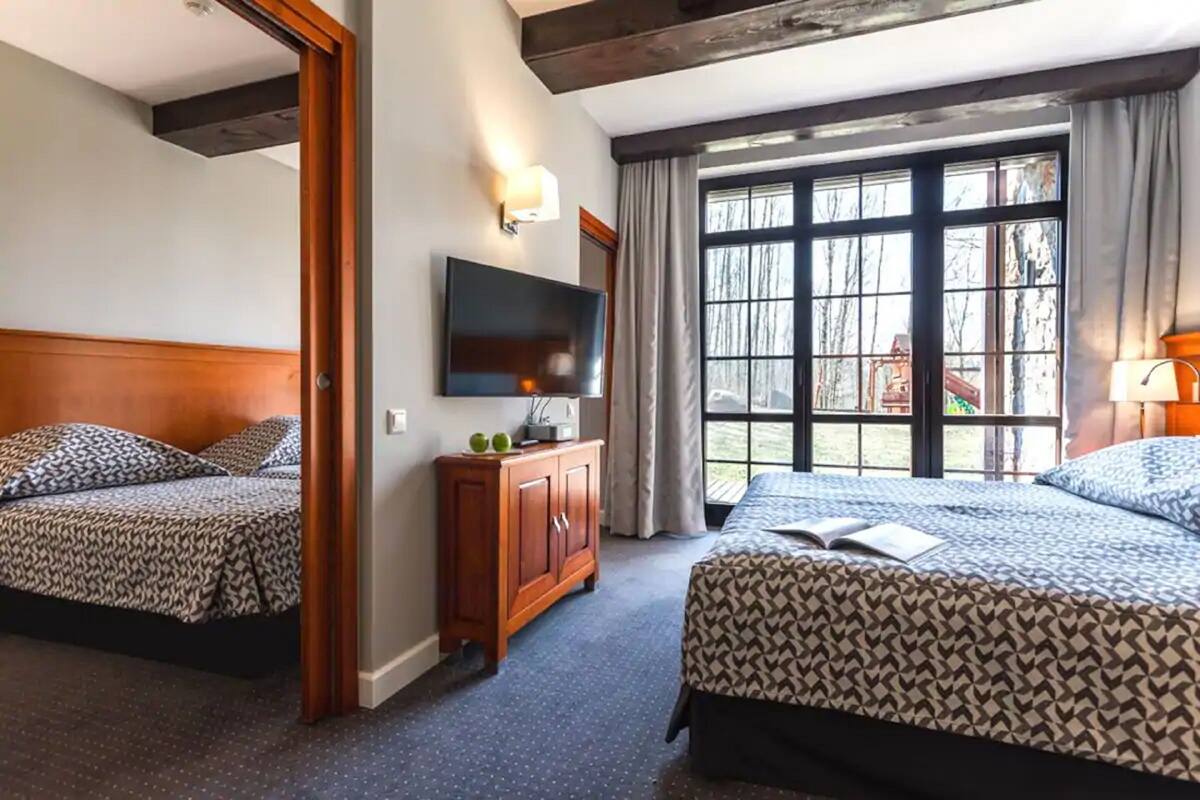 Habitación de hotel con dos camas, televisión, aparador de madera y ventanas amplias con cortinas.
