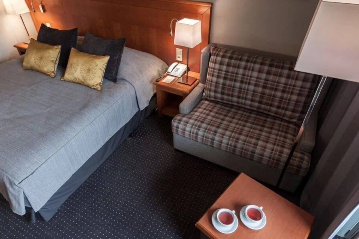 Una habitación de hotel con una cama, una silla, una mesita de noche con lámpara y una pequeña mesa con dos tazas.