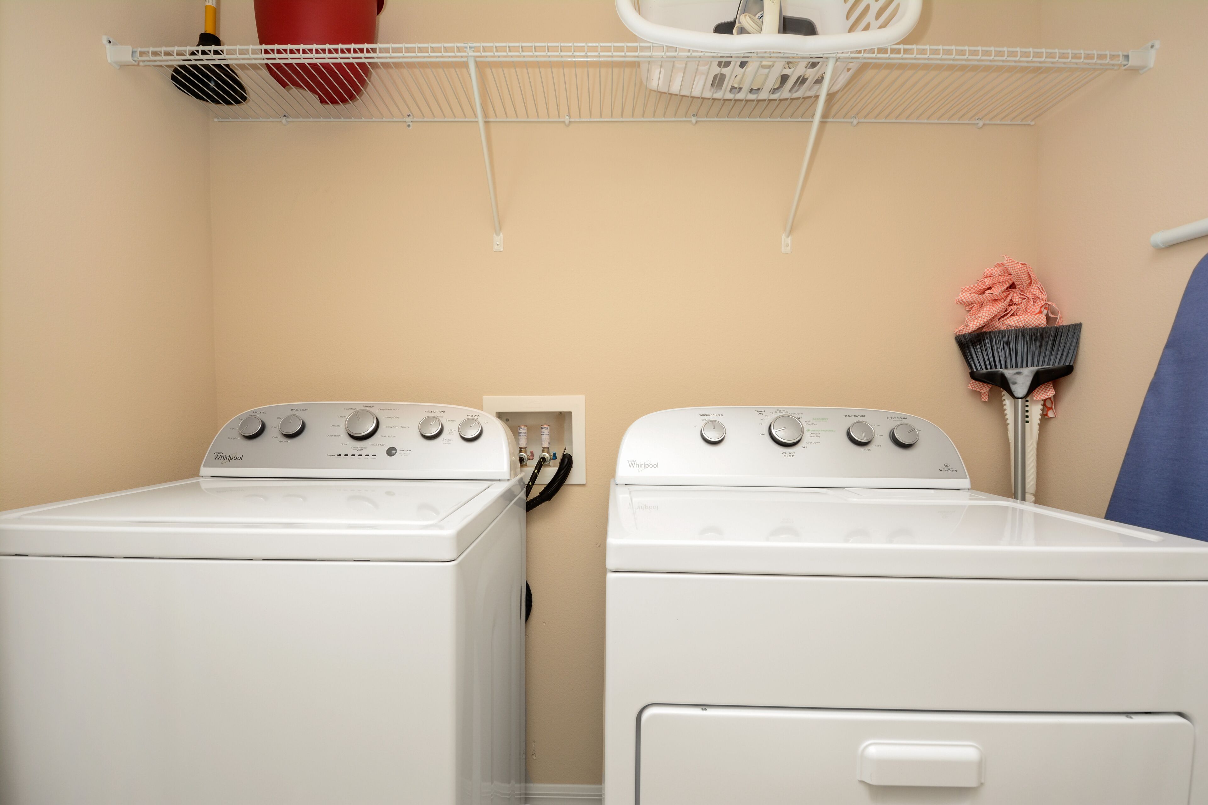 A laundry room with two white Whirlpool washers and dryers, a white shelf with cleaning supplies, and a blue curtain.