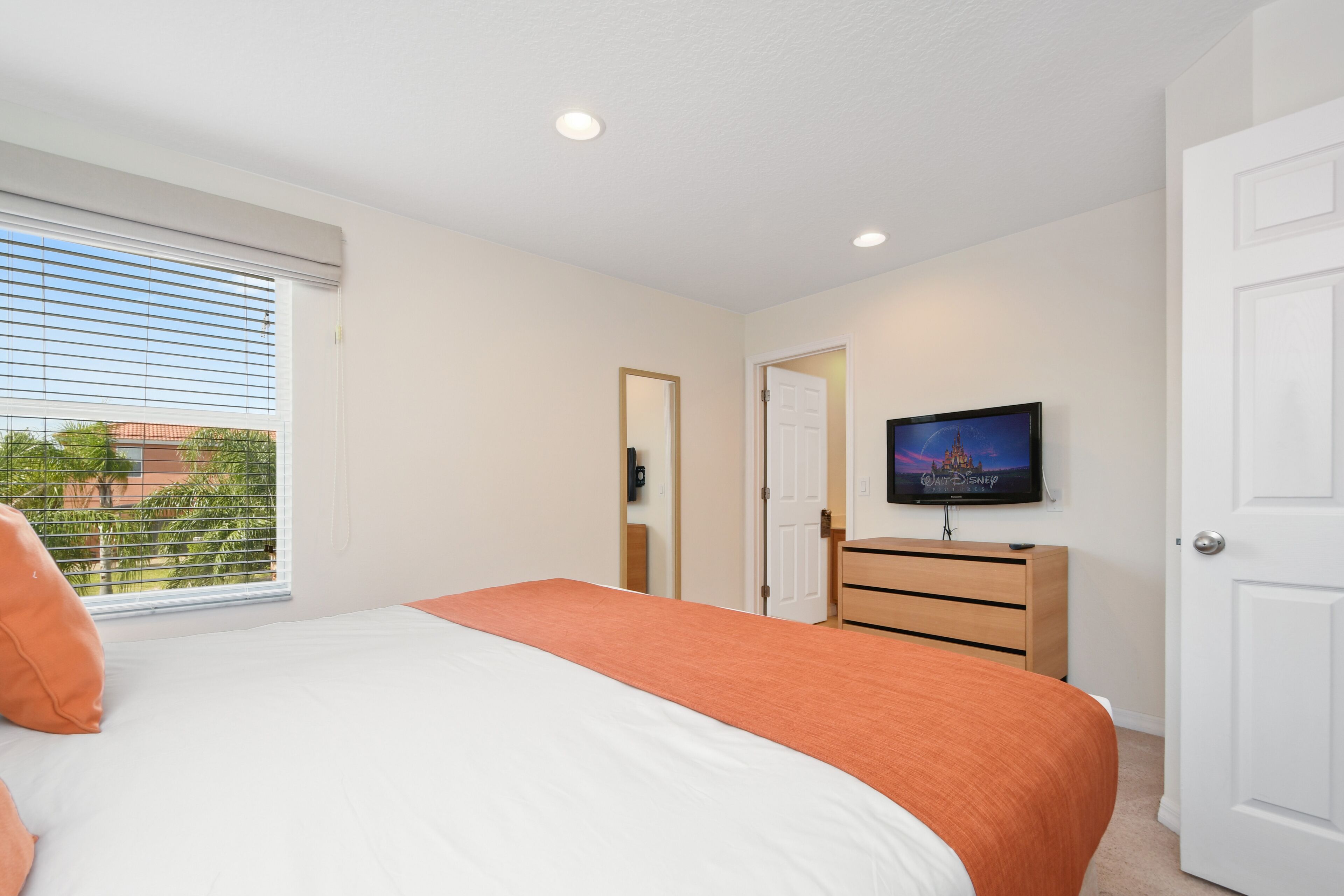 A bedroom with a bed, a dresser, a television on the wall, and a window with blinds.