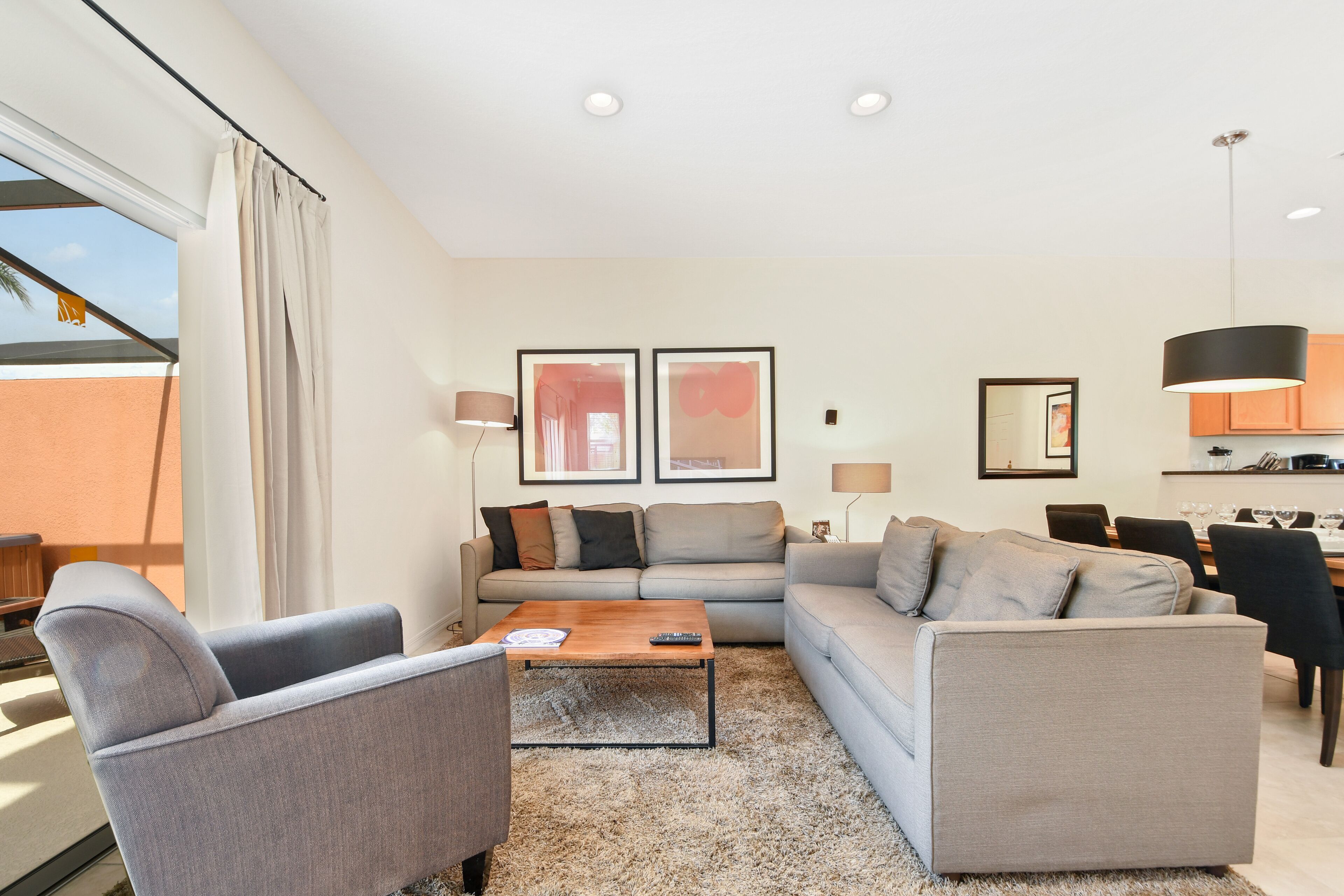 A modern living room with a sofa, armchair, and coffee table, a dining area with chairs and a table, and a kitchen with cabinets and a hanging light.