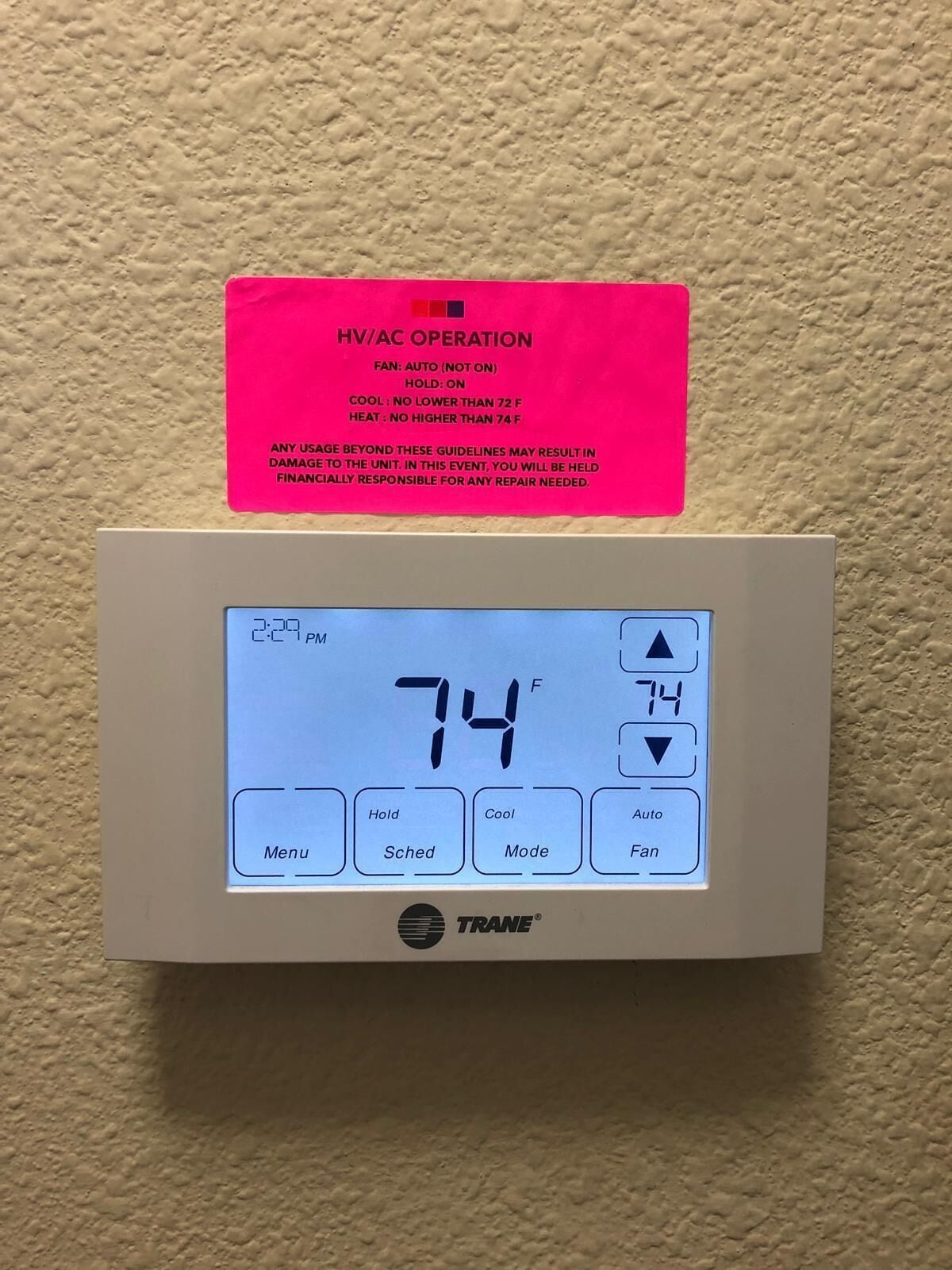 A TRANE HVAC control panel with a digital display showing 74°F, a pink maintenance tag, and operational buttons.