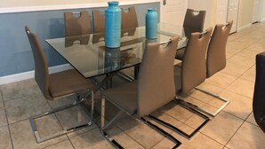 Uma sala de jantar com mesa de vidro e cadeiras marrons, um vaso azul e uma obra de arte emoldurada na parede.