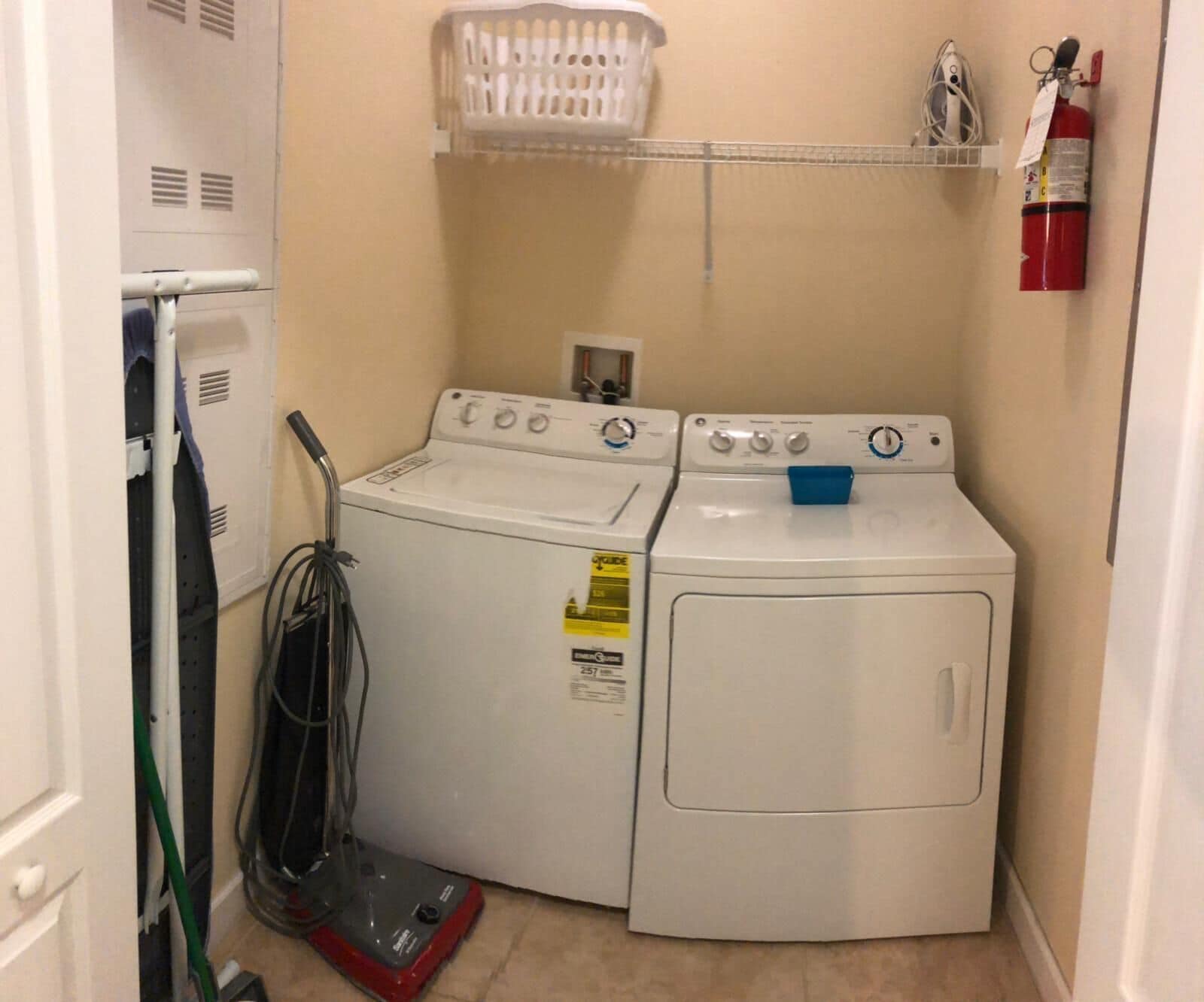 A laundry room with two washing machines, a dryer, a fire extinguisher, and a laundry basket on a shelf.