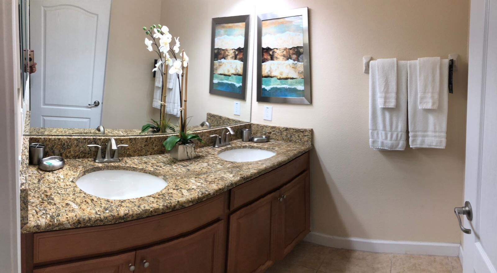 A bathroom with two sinks, granite countertops, wooden cabinets, a potted orchid, and two framed paintings on the wall.