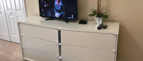 Un téléviseur posé sur un meuble blanc, avec une télécommande et une plante à côté.