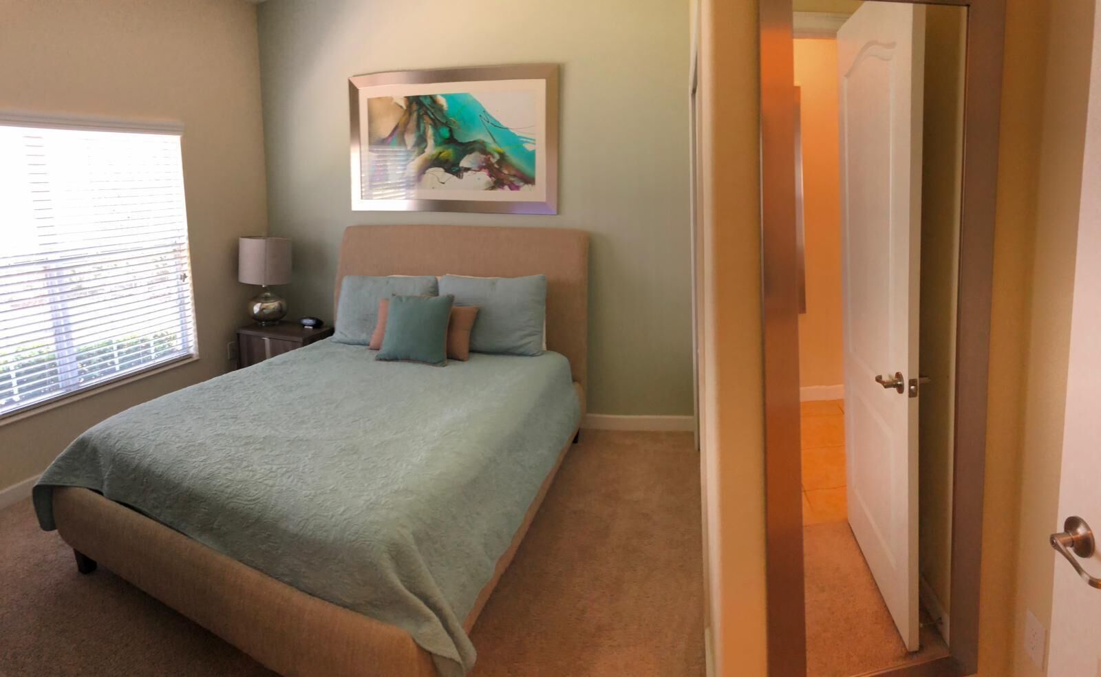 Un dormitorio con cama, mesita de noche, lámpara, un cuadro y una puerta.