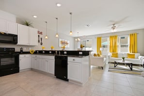 A modern kitchen with white cabinets, a black dishwasher, and a central island. There is a dining area with a white sofa and yellow curtains. - Lovely 4Bd Townhome Near Disney @ Compass Bay 5122 (Kissimmee)