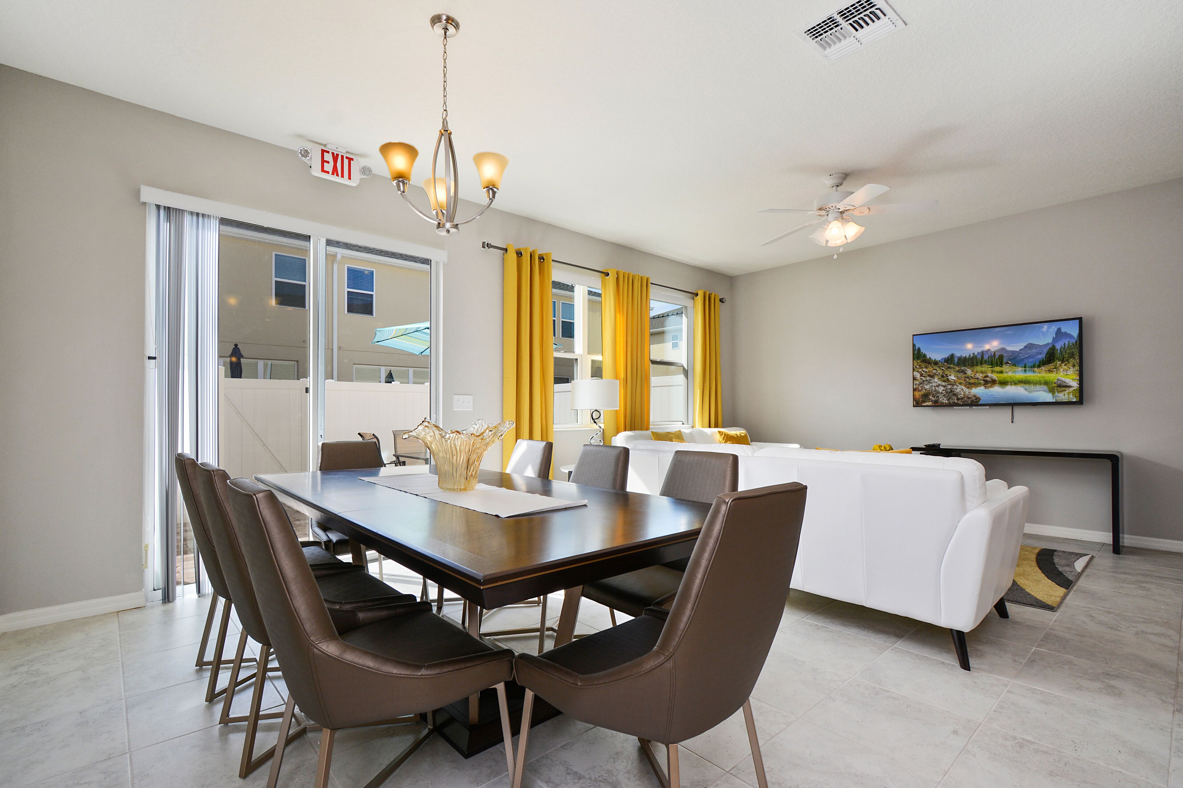 A modern dining area with a long table, chairs, and a chandelier. There is a sofa and a television in the background.