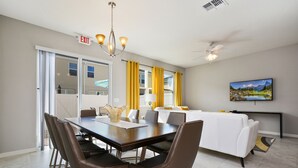 Uma sala de jantar moderna com uma mesa comprida, cadeiras e um candelabro. Há um sofá e uma televisão ao fundo.