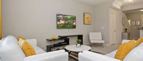 A modern living room with white sofas, a round coffee table, and a flat-screen TV mounted on the wall.